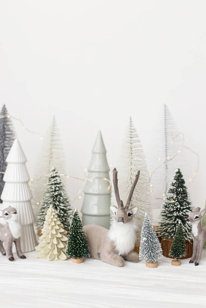 Κομψά χριστουγεννιάτικα δέντρα και παιχνίδια ταράνδων σε λευκό φόντο. Καλά Χριστούγεννα και καλές γιορτές! Χριστουγεννιάτικη σκηνή, μικροσκοπικό χιονισμένο δάσος. Σύγχρονη σκανδιναβική διακόσμηση - Φωτογραφία, εικόνα