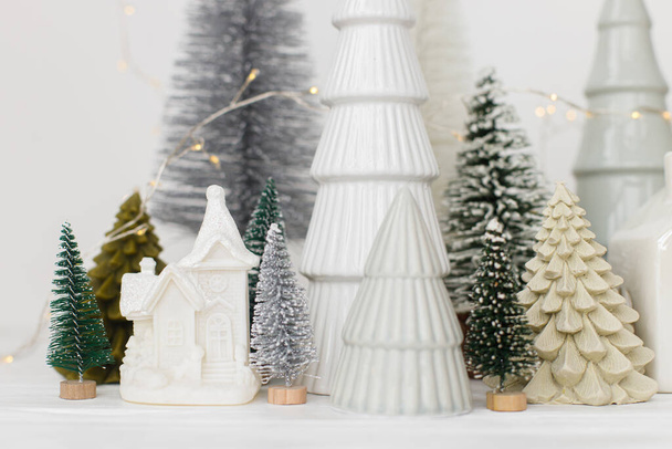Modern karácsonyi jelenet, miniatűr havas falu fehér asztalon. Boldog karácsonyt! Stílusos kis karácsonyfák és ház dekoráció. Téli ünnepi banner, skandináv monokromatikus dekoráció - Fotó, kép