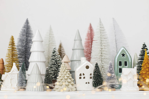 Merry Christmas! Stylish little Christmas trees and houses decorations on white table. Modern christmas scene, miniature cozy snowy village. Winter holiday banner, scandinavian decor - Photo, Image
