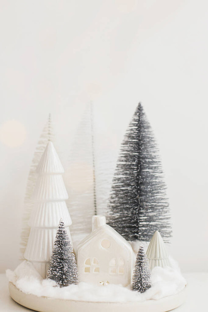 Zimowy hygge, przytulna scena świąteczna, miniaturowa śnieżna wioska na białym stole. Wesołych Świąt! Stylowe choinki i dom. Nowoczesny wakacyjny monochromatyczny wystrój, czas atmosferyczny - Zdjęcie, obraz