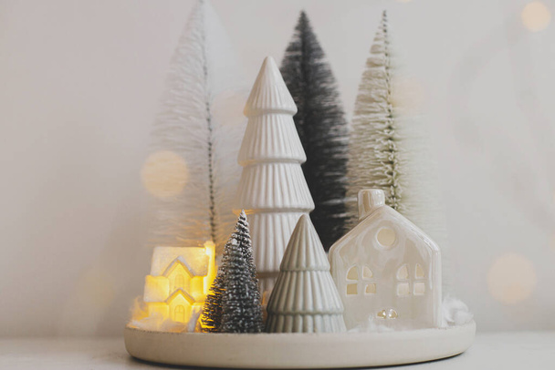 Χειμώνας hygge, ζεστό Χριστούγεννα μαγική σκηνή, μινιατούρα χιονισμένο χωριό με φώτα. Κομψά χριστουγεννιάτικα δέντρα και σπίτι σε λευκό τραπέζι. Καλά Χριστούγεννα! Μοντέρνα διακόσμηση διακοπών, ατμοσφαιρικός χρόνος - Φωτογραφία, εικόνα