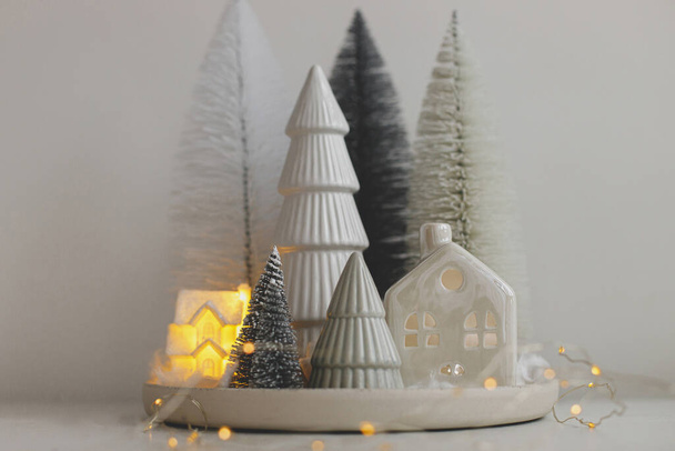 Buon Natale! Eleganti alberelli di Natale e casa su tavolo bianco. Inverno hygge, accogliente scena natalizia fata, villaggio nevoso in miniatura con luci. Decorazioni natalizie moderne - Foto, immagini