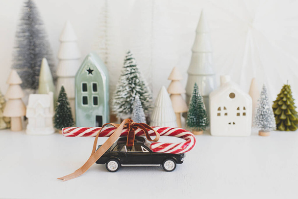 Merry Christmas and Happy Holidays! Stylish little car carrying candy cane on background of Christmas miniature snowy village. Festive winter scene on white table. Holidays are coming banner - Photo, Image