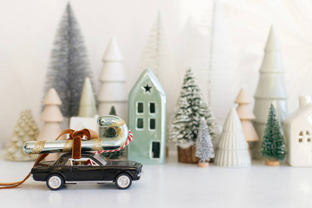 Elegante coche pequeño que lleva bastón de caramelo en el fondo de la aldea nevada miniatura de Navidad. Feliz Navidad y Felices Fiestas! Escena festiva de invierno sobre mesa blanca. Se acercan los días festivos - Foto, Imagen