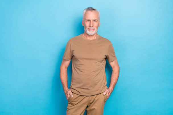 Photo of healthy handsome senior man with gray hairstyle stubble wear beige t-shirt hands in pockets isolated on blue color background. - Foto, Bild