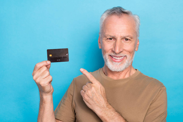 Portrait of cheerful good mood senior gray hair beard dressed beige t-shirt directing at plastic card isolated on blue color background. - Fotografie, Obrázek