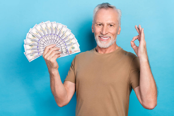 Portrait of satisfied retired senior with gray hair beard dressed beige t-shirt hold money showing okey isolated on blue color background. - Photo, Image