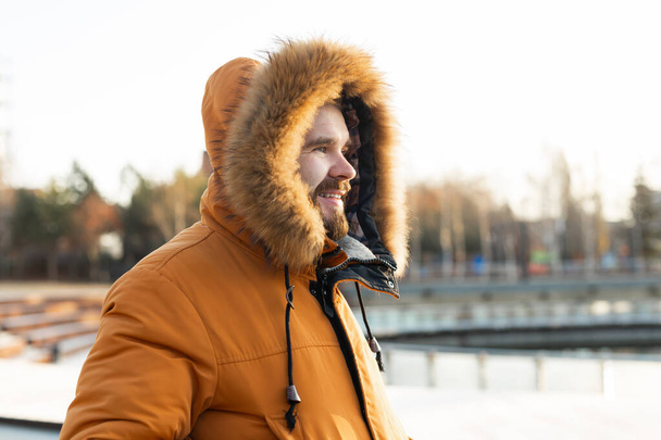 Açık hava kış portresi. Kışlık kıyafetler giymiş yakışıklı sakallı bir adam. Adam karlı bir parkta palto giyiyor. - Fotoğraf, Görsel