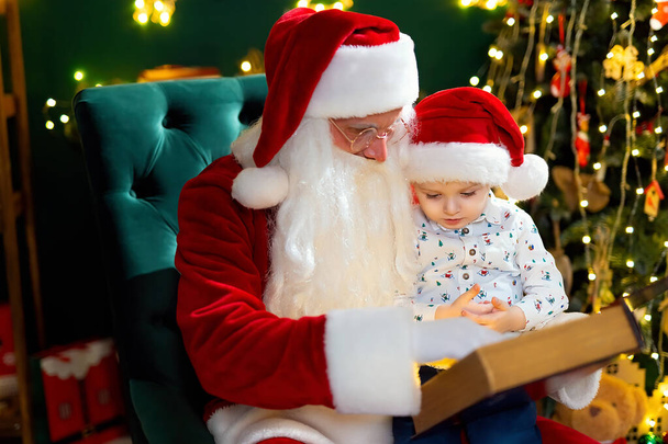 Santa Clause reads a fairytale to a little boy in Santas hat while sitting near Christmas tree. New Year concept. Christmas time  - Photo, Image