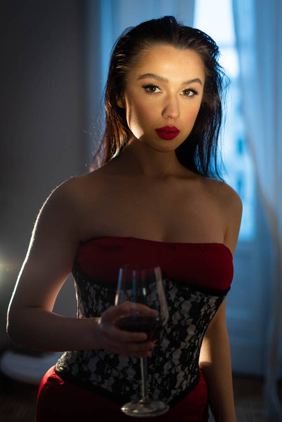 茶色の目と黒い髪をした美しい少女が、赤のドレスにプリント入りのコルセットを着て、ワインのグラスを手に持っている。白い窓を背景に。カメラを見ると肖像画. - 写真・画像