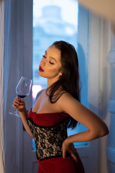Egy fényűző nő fényes piros ruhában, fűzővel az ablak mellett áll egy pohár borral. Romantikus vacsora. A luxusnak nincsenek korlátai, oldalra néz.. - Fotó, kép
