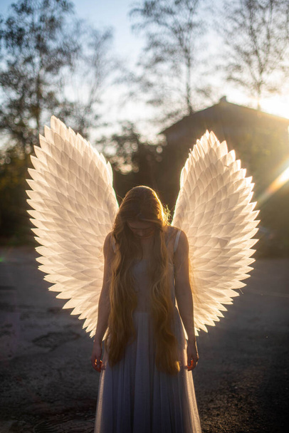 Young gentle angel girl stands in front of the autumn sun. Her white wings glow with beauty. She has long blond hair. She stands with her head and hands down. Portrait. - Photo, Image