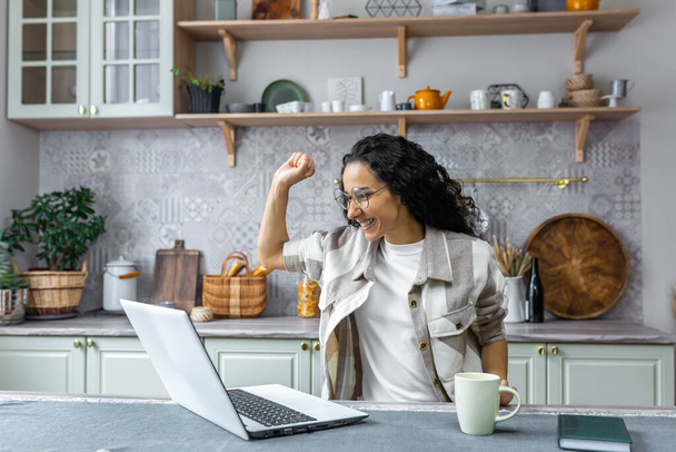 Hispanic business woman working remotely from home freelancer celebrating victory and success holding hands up looking at laptop reading good news. - Φωτογραφία, εικόνα