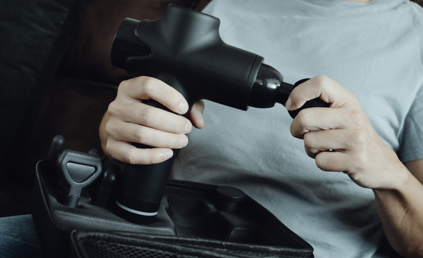 closeup of a young caucasian man, sitting on a brown couch, about to change the accessory of a massage gun, in a panoramic format to use as web banner or header - Photo, Image