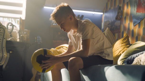 Mladý chlapec drží v rukou žlutý míček. Myslí na svůj zápas. Sedí na gauči v jeho ložnici. Vážné výrazy. Fotbalový zápas, sportovní koncept. - Fotografie, Obrázek