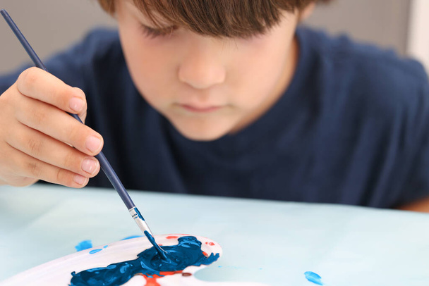 Close up of a little kid that mixes colors with a drawing brush. There is a palette with blue and red paint on the table. The boy is sitting outside, wearing blue T-shirt and looking at the paints - Photo, image
