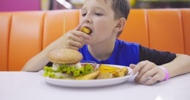 A gyerek hamburgert és sült krumplit eszik egy gyorsétteremben. A gyerek sült chipset mártott paradicsomszószba. Kiváló minőségű 4k felvételek - Felvétel, videó