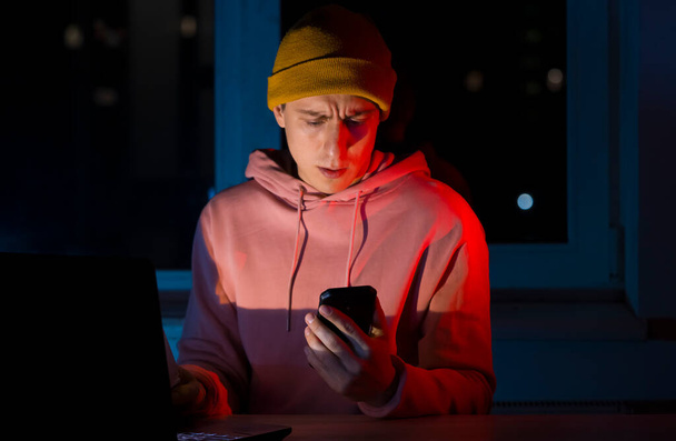 Een jongeman werkt 's avonds laat op een laptop thuis en praat over de telefoon, lost zakelijke problemen op, analyseert bestanden, freelancer communiceert online. - Foto, afbeelding