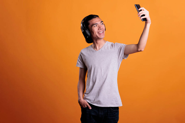 Lachende Aziatische tiener het nemen van foto 's op smartphone camera aan de voorkant. Jonge vrolijke aantrekkelijke man in draadloze hoofdtelefoon met mobiele telefoon, poseren voor selfie, studio medium shot - Foto, afbeelding