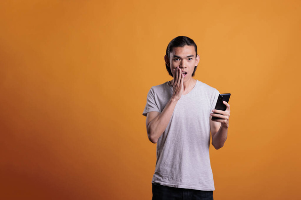 Shocked young asian man looking at smartphone, reading news online, medium shot. Teenager standing with open mouth and hand on face, holding mobile phone, hearing gossips - Φωτογραφία, εικόνα