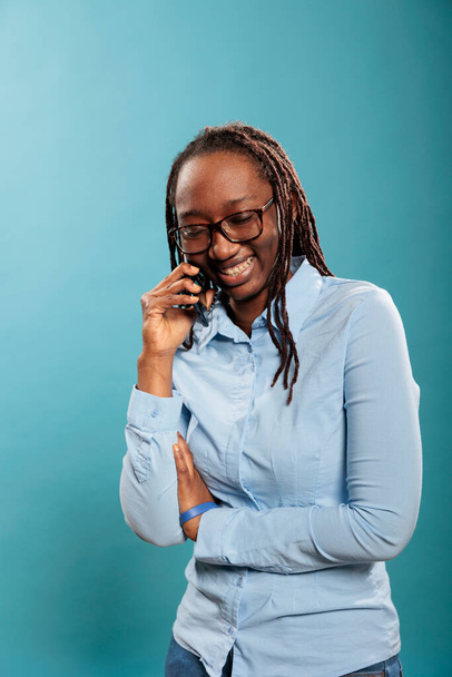 Cheerful adult person talking with friend on touchscreen cellphone device. Happy joyful young woman smiling heartily while having a phone call conversation on blue background. Studio shot - Photo, image