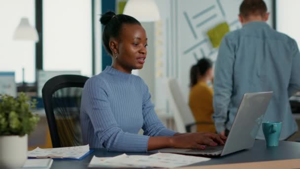 Portrait of african american woman typing using laptop keyboard and smiling at camera sitting at desk in busy startup office. Casual business employee working relaxed with sales statistics. - Filmagem, Vídeo