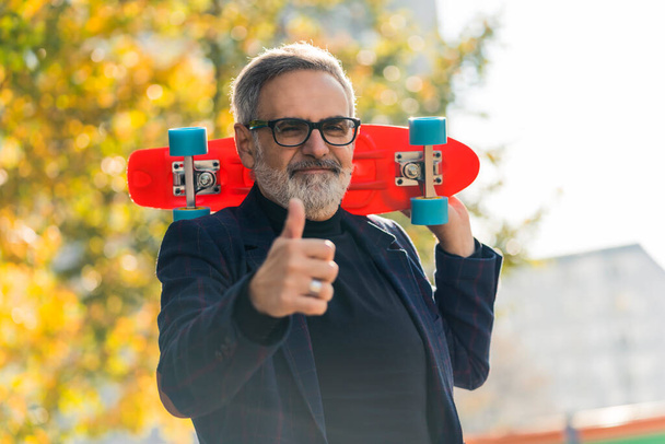 Grey-haired middle-aged man in glasses wearing blazer holding colorful skateboard and showing thumbs up to camera smiling. Silly grandpa. Horizontal outdoor shot. High quality photo - Фото, зображення