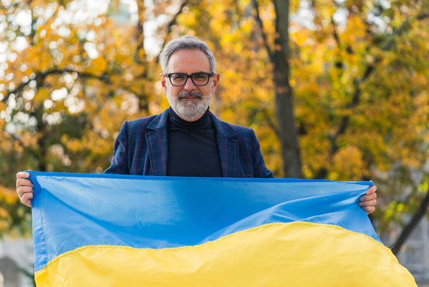 Hombre de mediana edad de pelo gris parado en el parque de la ciudad sosteniendo la bandera ucraniana mirando a la cámara sonriendo. Apoyo a los ucranianos. Protesta. Tiro horizontal al aire libre. Foto de alta calidad - Foto, Imagen