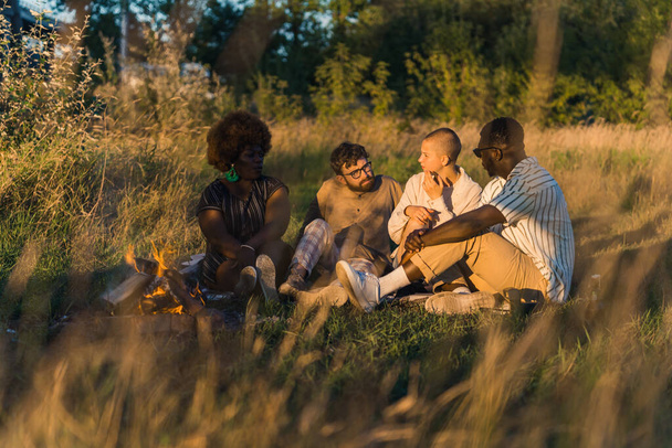 自然の中で人々の美しいシーン。多人種の友人- 2人の男性と2人の女性-彼らの前に少したき火をして芝生の上に座っている。屋外パーティー中のカジュアルな会話。高品質 - 写真・画像
