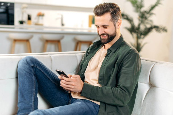 Positive handsome stylish caucasian man sits on a sofa in a cozy living room, uses his smartphone, spends leisure time online, chats with friends on social networks, browsing the internet, smiles - Fotó, kép