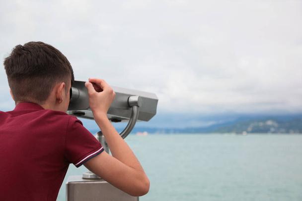 Teenage boy looking through mounted binoculars at mountains. Space for text - Фото, изображение