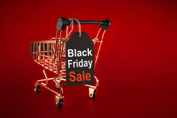 Μαύρη ετικέτα πώλησης Παρασκευή και χρυσό καλάθι αγορών πάνω από το κόκκινο φόντο με αντίγραφο χώρο για κείμενο. Μαύρη Παρασκευή πώληση, έννοια ψώνια - Φωτογραφία, εικόνα