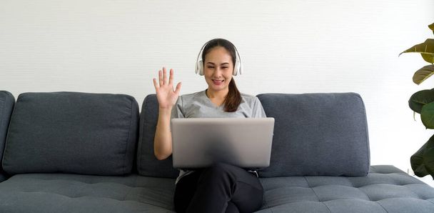 Azjatka w szarej koszulce i słuchawce machająca ręką przez zdalną komunikację przez laptop w salonie.  - Zdjęcie, obraz