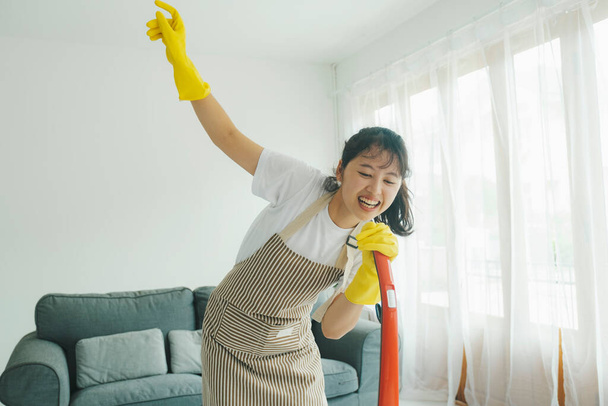 Cheerful young woman having fun singing while cleaning using vacuum cleaner at home. Woman in casual clothes and apron cleaning, singing, dancing, and having fun while cleaning. Housekeeping concept. - Foto, imagen