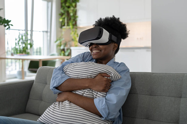 VRヘッドセットを持つ楽観的なアフリカ系アメリカ人女性は恐怖と喜びを抱き枕を笑顔。素晴らしいです黒の女の子テスト現代的なハイテクヘルメット座っています上のソファ見映画とともに仮想現実 - 写真・画像