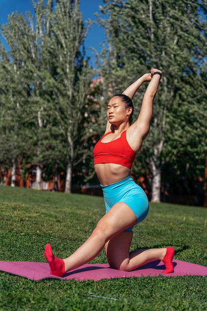 Young asian woman wearing sports clothes stretching in her mat in the park. She is concentrated and looking at front. - Foto, imagen