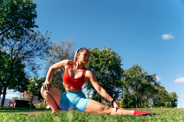 Focused asian woman wearing sports clothes stretching in her mat in the park. She is concentrated and looking at front. - Fotó, kép