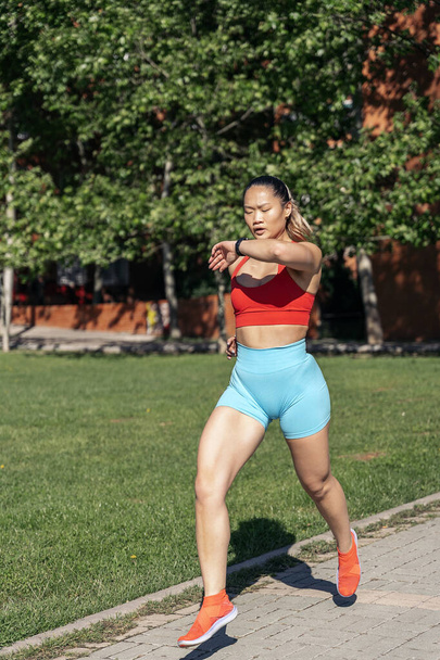 Active asian woman with sports clothes running in the park during sunny day. She is checking her smart-watch. - Photo, Image