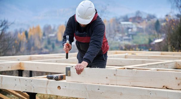 Man bouwend houten frame huis op paalfundering. Timmerman installeert meubels voor houten planken, met behulp van hamer en schroevendraaier. Timmermansconcept. - Foto, afbeelding