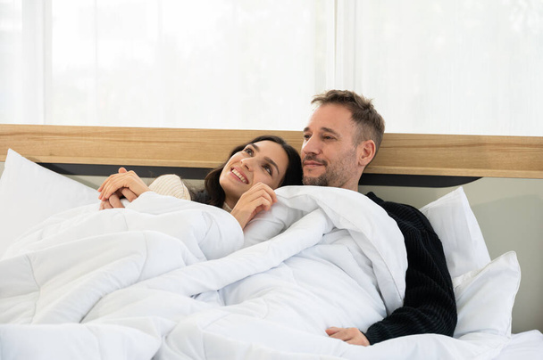 Heureux couple souriant homme et femme dans des draps chauds et une couverture embrassant au lit, parlant et s'amusant ensemble tandis que la femme appuyait sa tête sur son épaule et appréciait profiter d'un matin d'hiver paresseux à la maison - Photo, image