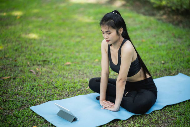 Asian sport fitness woman learning by doing a yoga according to a video clip or  video conferrence with teacher. Asian woman using tablet and doing yoga in park. Yoga study learning online concept. - Photo, Image