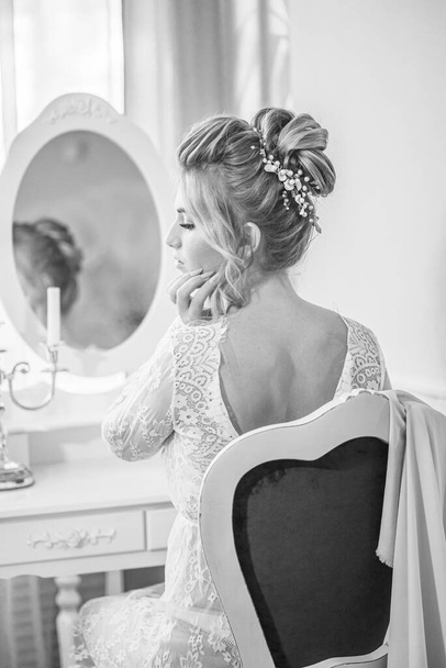 A young beautiful bride with a wedding hairstyle and makeup in the morning in a floor-length white lace tunic near the boudoir table. wedding morning - Photo, image