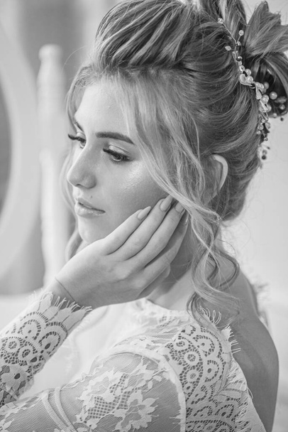 Eine junge schöne Braut mit Hochzeitsfrisur und Make-up am Morgen in einer bodenlangen weißen Spitzenjacke in der Nähe des Boudoir-Tisches. Hochzeitstag - Foto, Bild