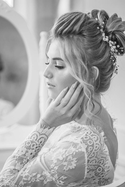 A young beautiful bride with a wedding hairstyle and makeup in the morning in a floor-length white lace tunic near the boudoir table. wedding morning - Foto, immagini