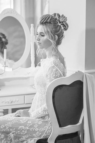 A young beautiful bride with a wedding hairstyle and makeup in the morning in a floor-length white lace tunic near the boudoir table. wedding morning - Fotó, kép