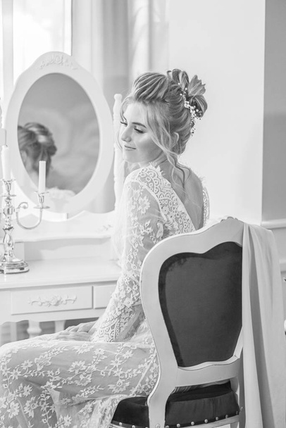 A young beautiful bride with a wedding hairstyle and makeup in the morning in a floor-length white lace tunic near the boudoir table. wedding morning - Valokuva, kuva