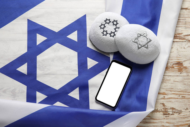 Flaga Izraela, kippah i telefon komórkowy na jasnym drewnianym tle - Zdjęcie, obraz