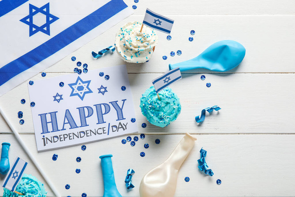 Kaart met tekst HAPPY INDEPENDENCE DAG, vlag van Israël, cupcakes en feestelijke decor op witte houten achtergrond, close-up - Foto, afbeelding