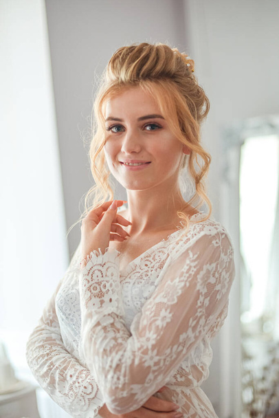 A young beautiful bride with a wedding hairstyle and makeup in the morning in a floor-length white lace tunic near the boudoir table. wedding morning - Foto, imagen