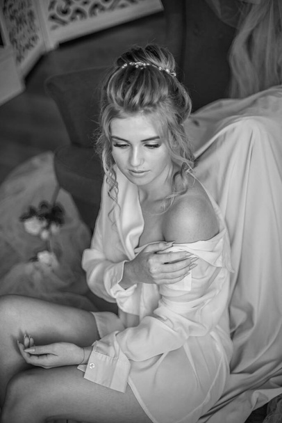 Young beautiful bride with wedding hairdo and makeup in the morning wearing a white silk negligee in the bedroom. wedding morning - Zdjęcie, obraz